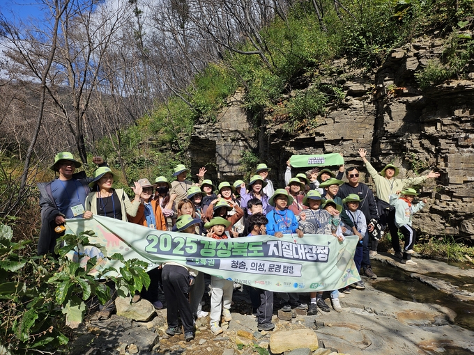 ▲ 참가자들이 25 경북 의성군 송내리 공룡발자국 화석산지에서 진행된 ‘2025 경상북도 내륙권 지질대장정’ 의성 구간 탐방 중 기념 현수막을 펼치고 복원된 산림을 배경으로 손을 들어 인사하고 있다. 의성군
