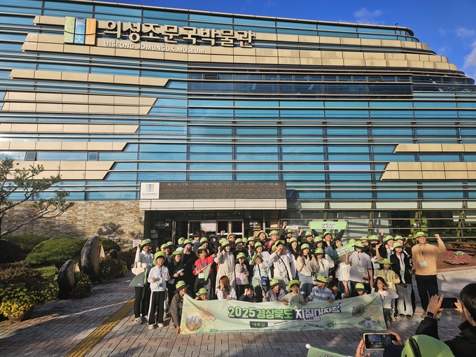 ▲ 참가자들이 24일 경북 의성군 의성조문국박물관 앞에서 ‘2025 경상북도 내륙권 지질대장정’ 의성 구간 출발에 앞서 단체로 기념 현수막을 들고 박물관 전면 유리 외벽을 배경으로 환하게 손을 들어 인사하고 있다. 의성군