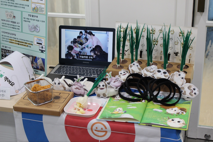 ▲ 의성군어린이사회복지급식관리지원센터가 ‘제14차 식생활교육매체 경진대회’에 출품한 유아용 식생활교육 교재·교구 ‘육쪽마늘 쪽쪽쪽’ 전시물이 24일 서울 서초구 aT센터 현장 부스에 전시돼 있으며, 마늘 캐릭터 교구·동화책·영상자료 등이 함께 배치되어 있다. 의성군어린이사회복지급식관리지원센터