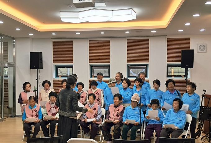 ▲ 오는 12월 20일 ‘인생 시 낭독’ 무대를 앞두고 의성군청년센터 강당에서 산불 피해 어르신들이 파란 단체복과 분홍 조끼를 차려입은 채 지도 강사의 동작을 따라 시 구절을 맞춰 읽어 나가며, 잃어버린 일상 위에 다시 목소리로 삶을 세우는 장면이 따뜻하게 담겼다. 의성군